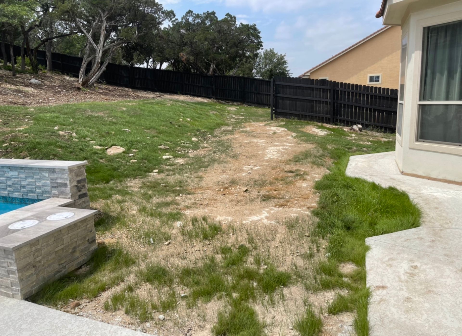 Backyard with patchy grass, a pool edge, concrete patio, and black fence by a house