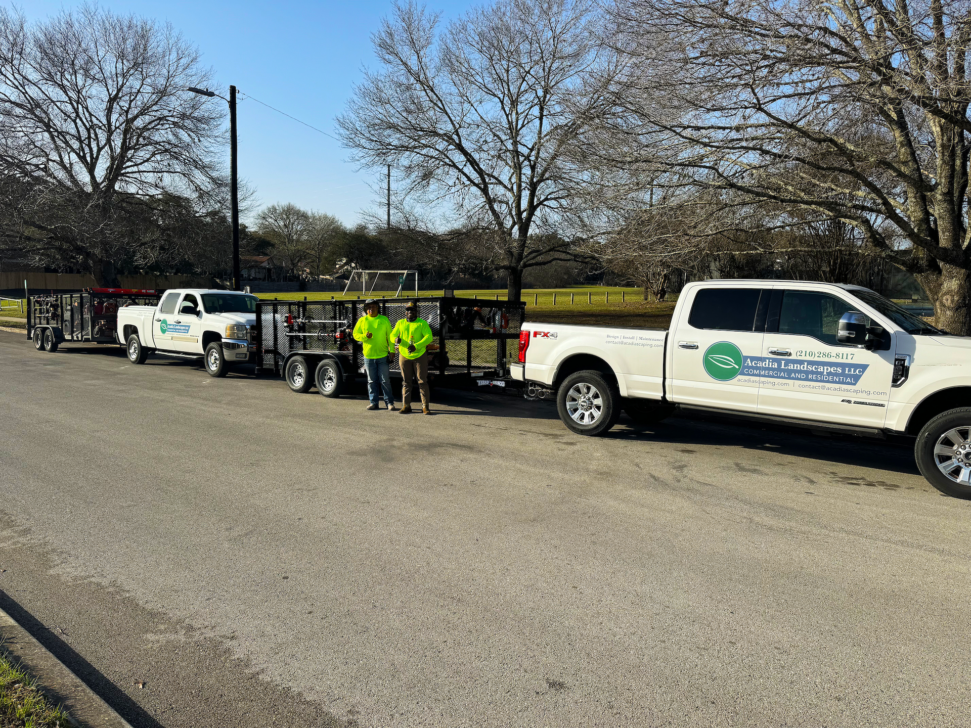 Utility crew beside white pickup trucks and equipment trailer on a roadside under bare trees.
