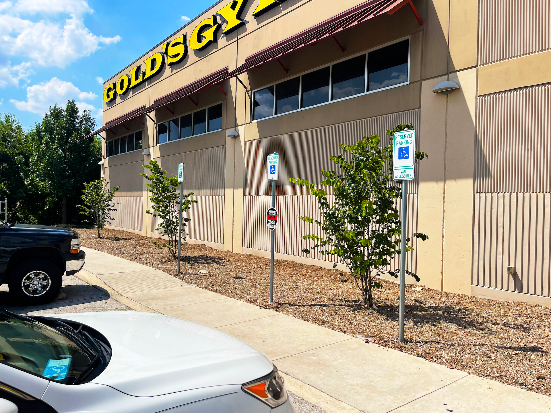 Exterior of Gold's Gym building with accessible parking signs and parked cars on a sunny day.