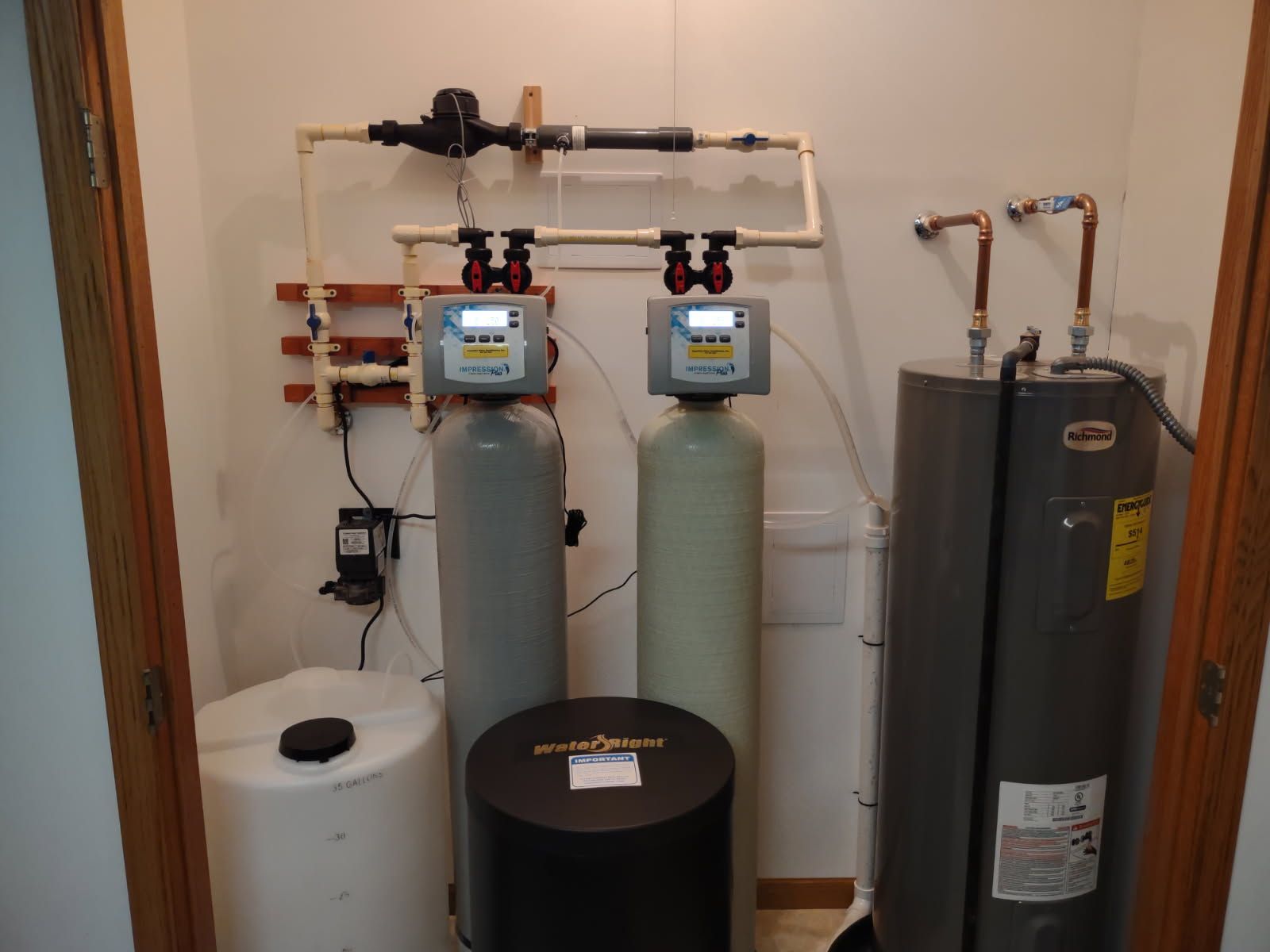 Water softener and filtration system in a utility room, with two gray tanks and a black brine tank.