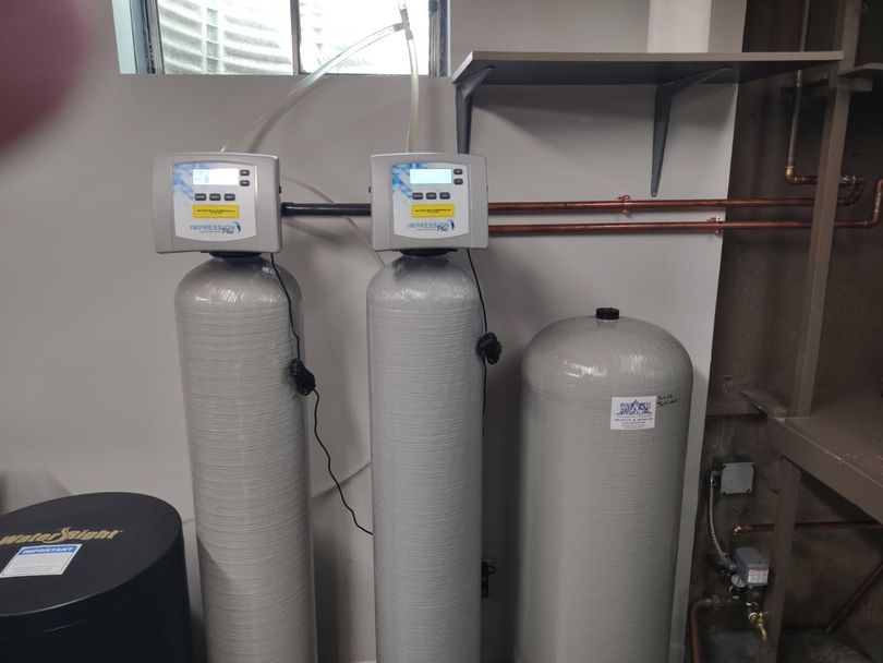 Two gray water softener tanks and a resin tank connected by pipes in a utility room.