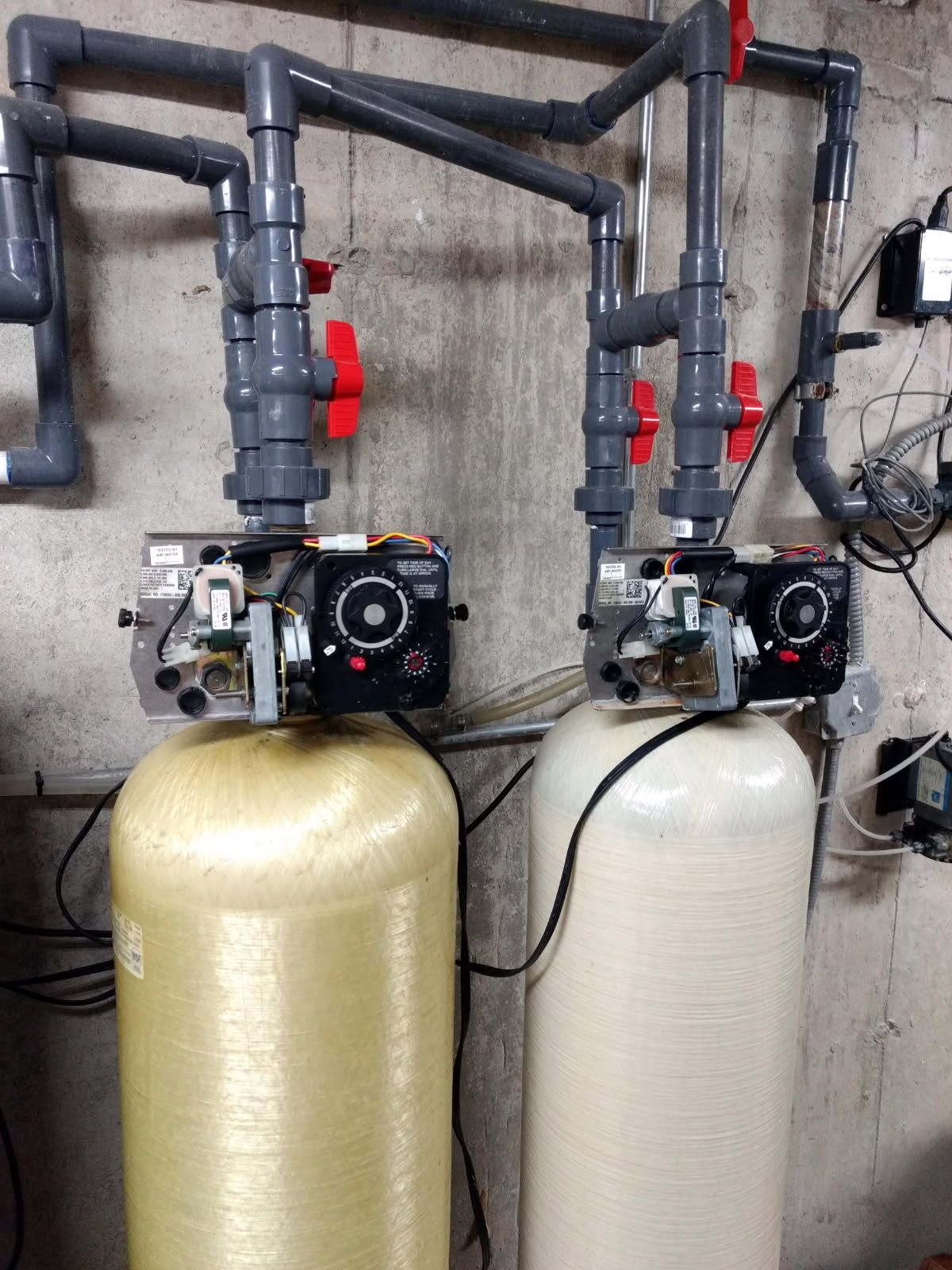 Two water softener tanks with black control valves and black piping in a utility room
