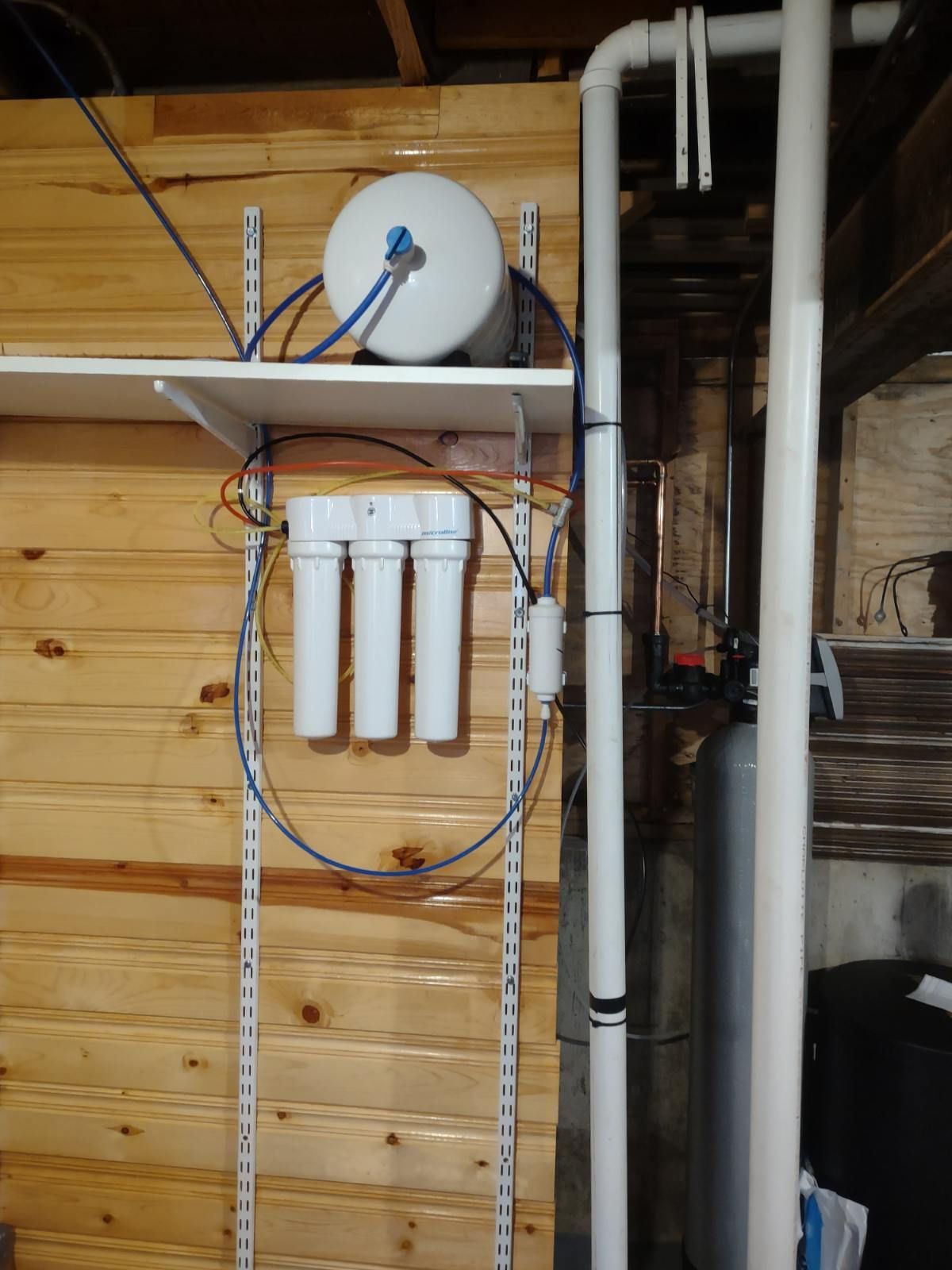 Wall-mounted white water filter system with hoses and tubing in a wood-paneled basement utility area