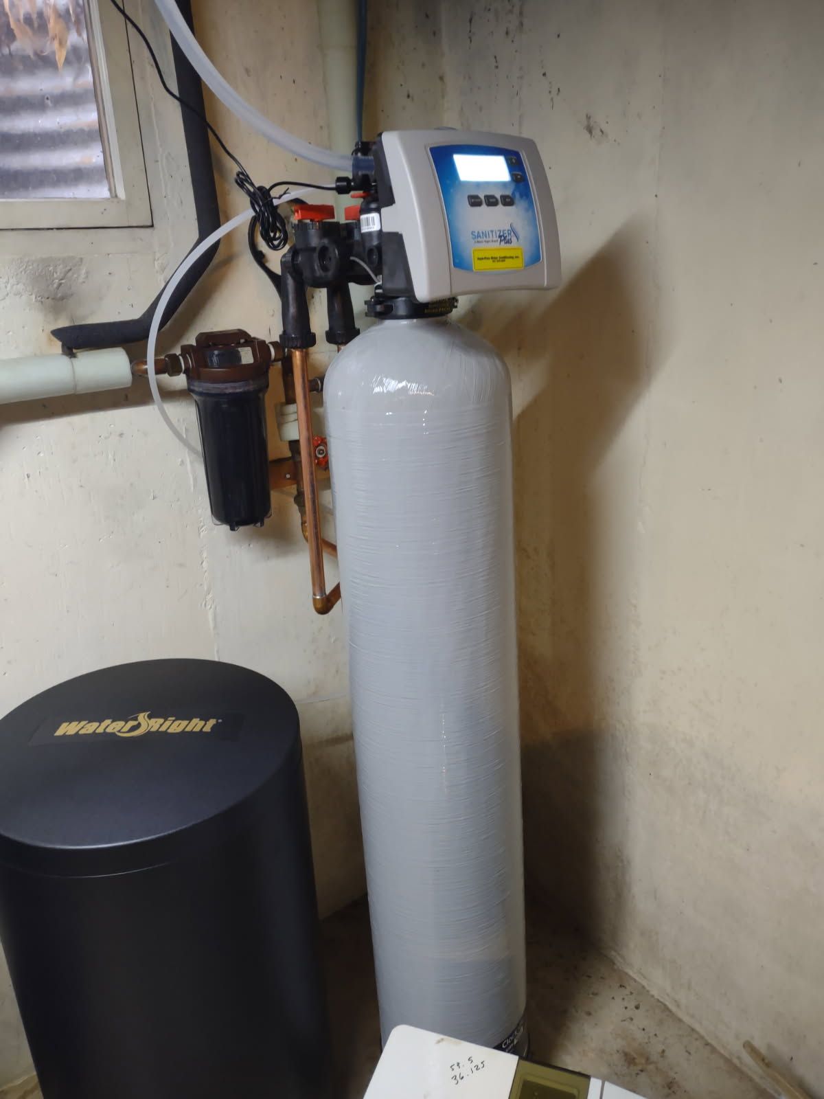 Water softener system with gray tank, black filter, and blue control head in a utility room corner