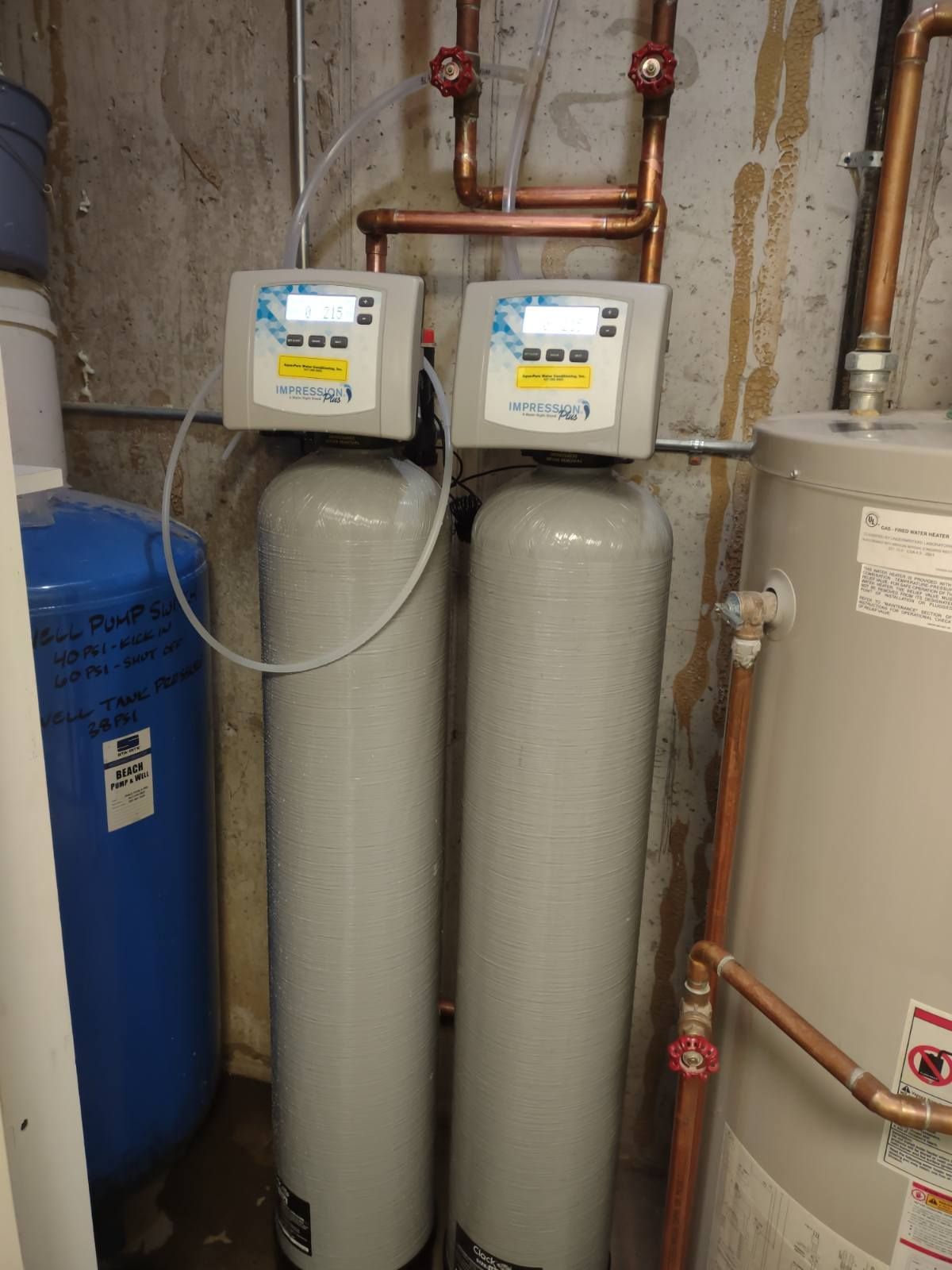 Two tall beige water softener tanks beside a blue tank and pipes in a utility room