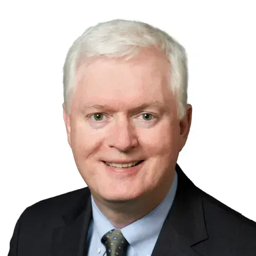 Portrait of a smiling man in a dark suit, light blue shirt, and patterned tie on a white background