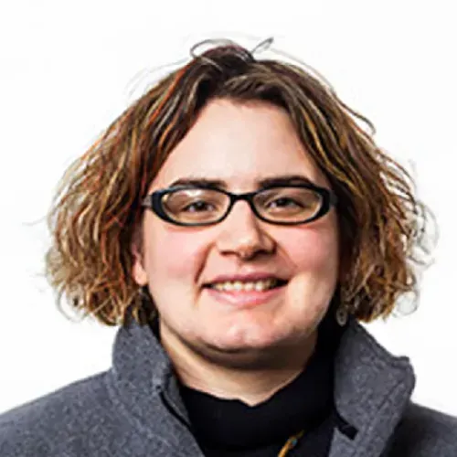 Person with glasses and curly hair smiling in a gray coat against a white background