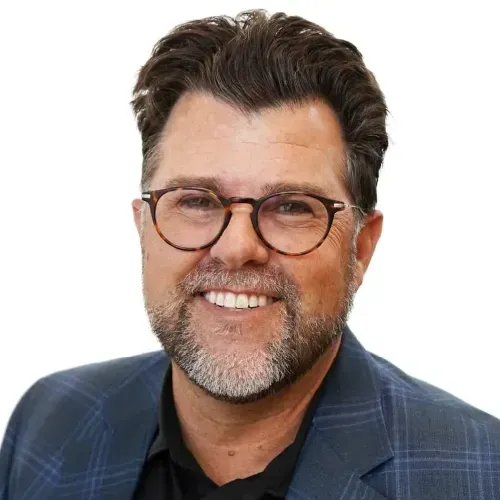 Smiling man with glasses and salt-and-pepper beard in a dark plaid blazer on a white background
