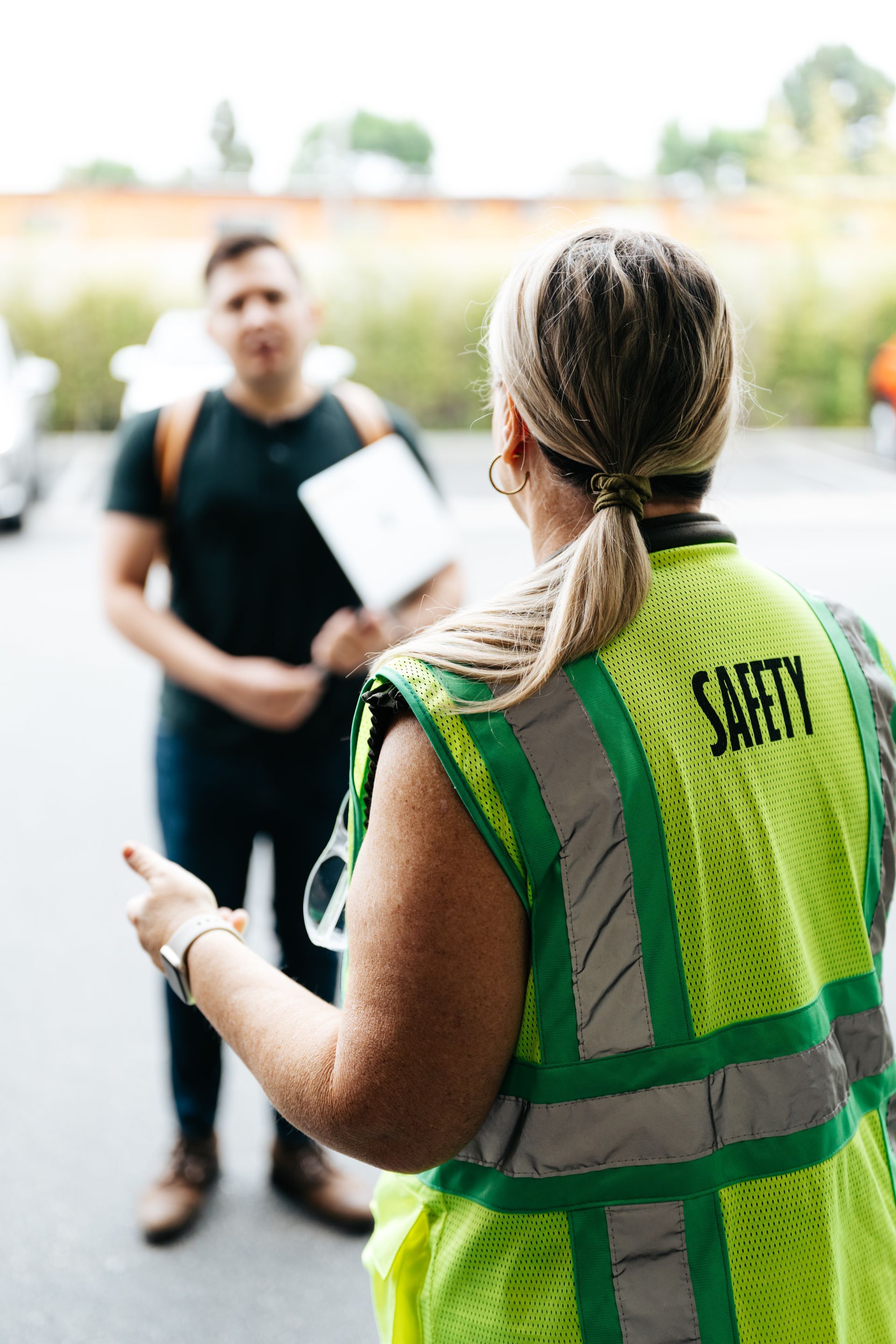 Safety vest-clad worker speaking to a person outdoors, holding a clipboard and paperwork.