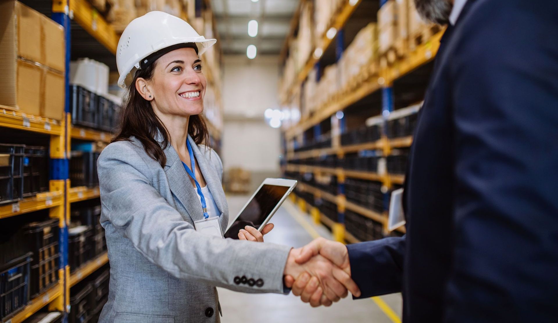 Warehouse worker in a hard hat shaking hands while holding a tablet in an aisle of stacked shelves