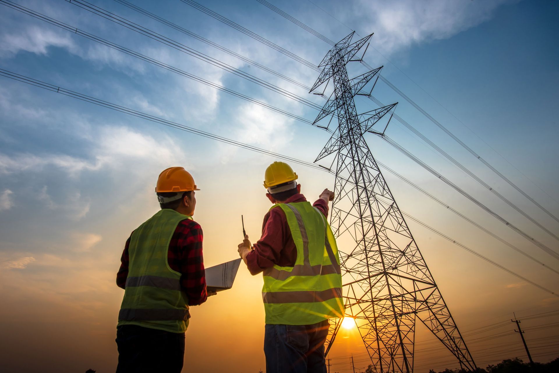 Two utility workers in hard hats inspect power lines at sunset beside a transmission tower.