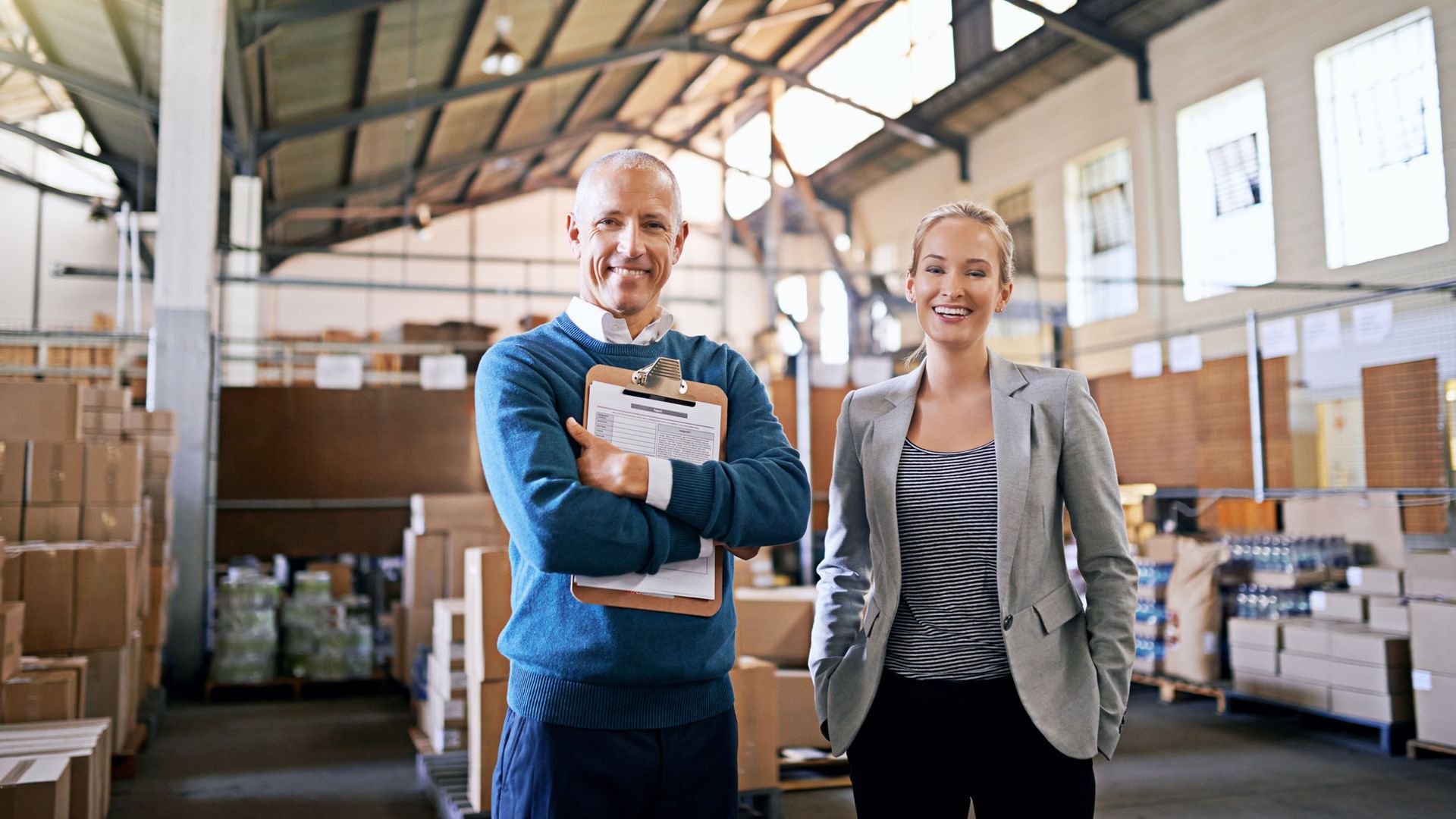 Two warehouse workers smiling in a storage facility, one holding a clipboard with boxes and pallets behind them