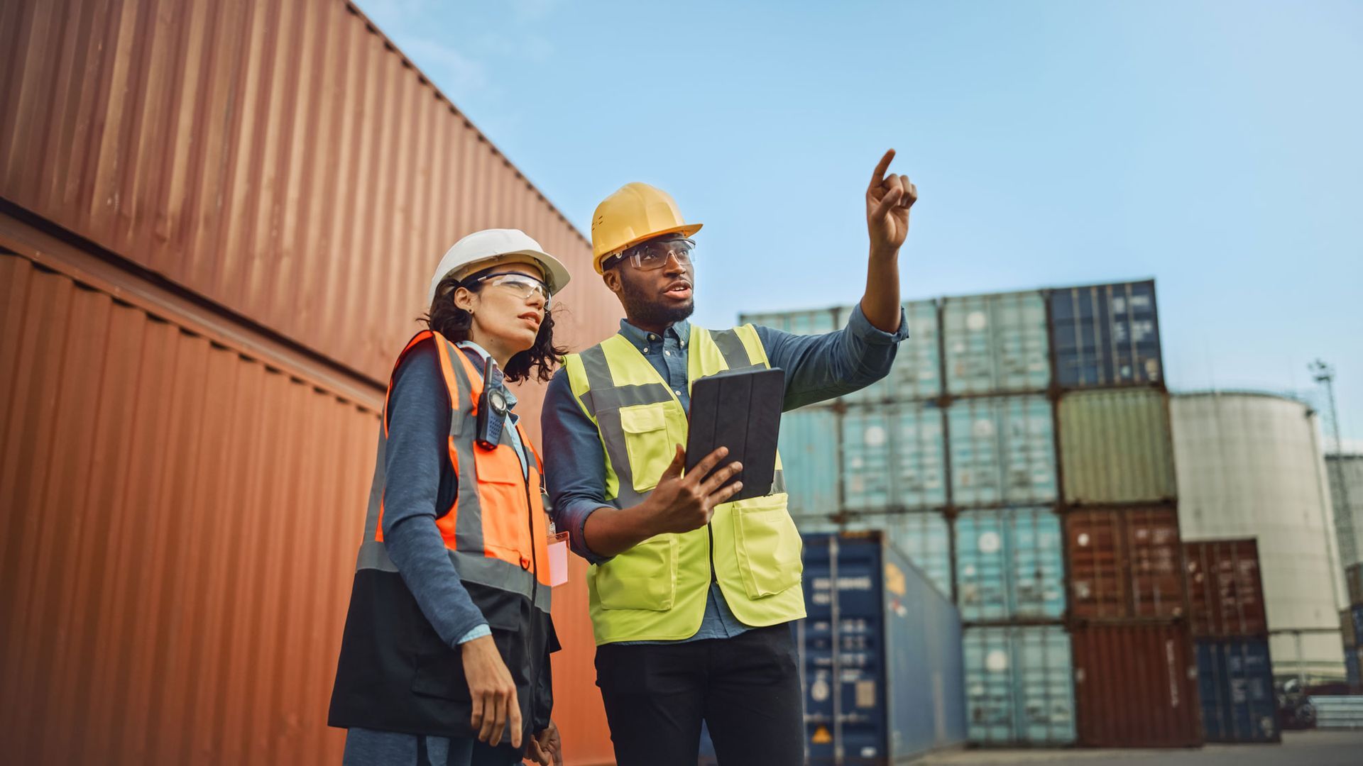 Two workers in safety vests and hard hats inspect shipping containers at a port, one pointing ahead.