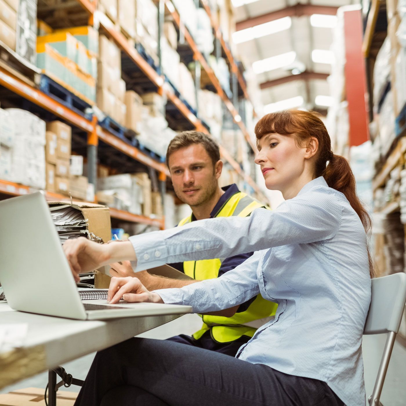 Two warehouse workers in safety vests use a laptop at a desk in an aisle.