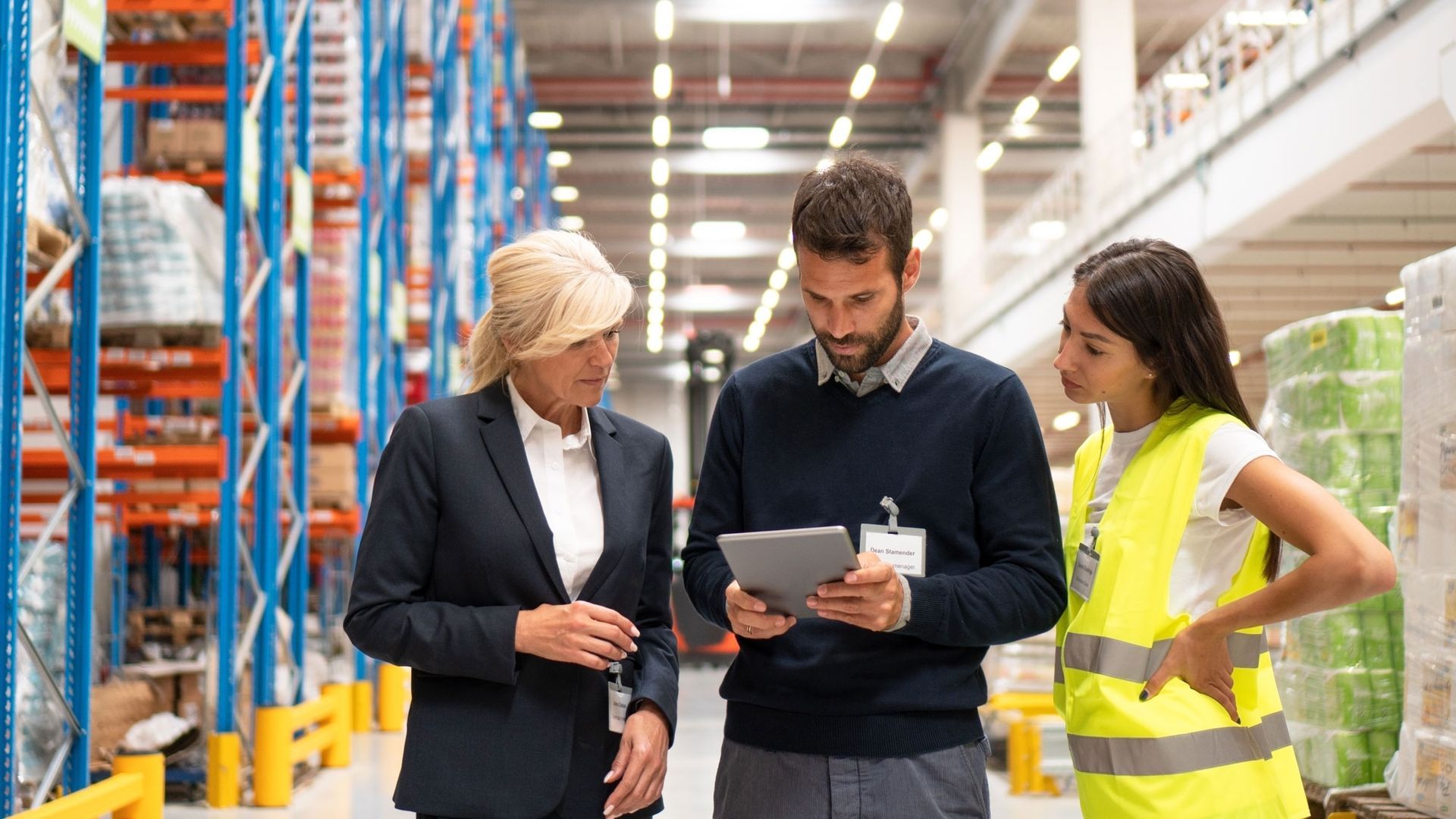 Three warehouse workers review a tablet in an industrial aisle, with one wearing a yellow safety vest