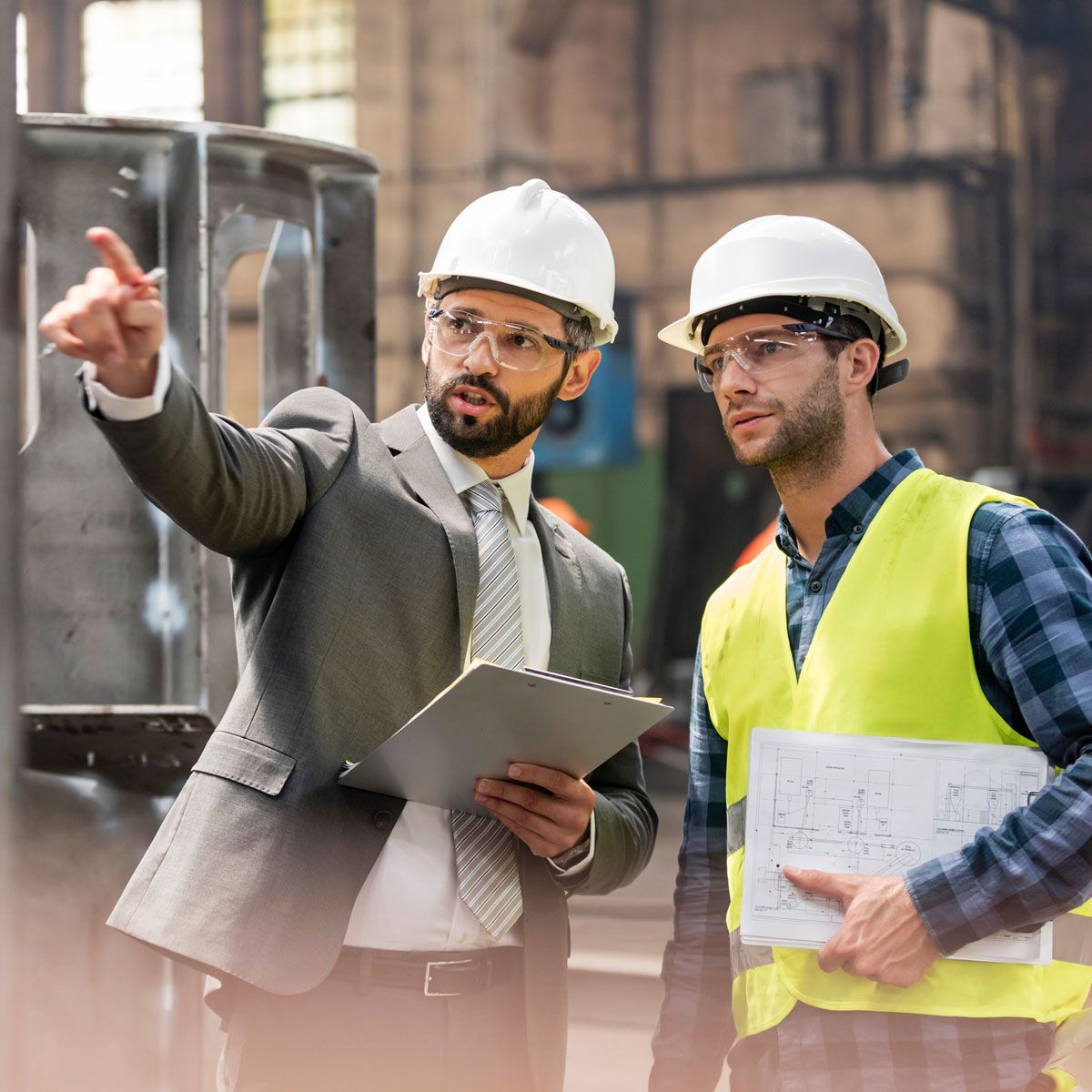 Two construction workers in hard hats review plans at an industrial site, one pointing ahead.