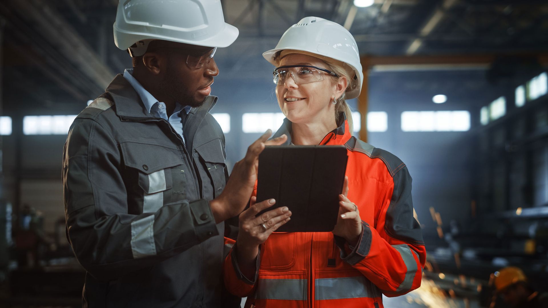 Two workers in hard hats reviewing a tablet in an industrial warehouse