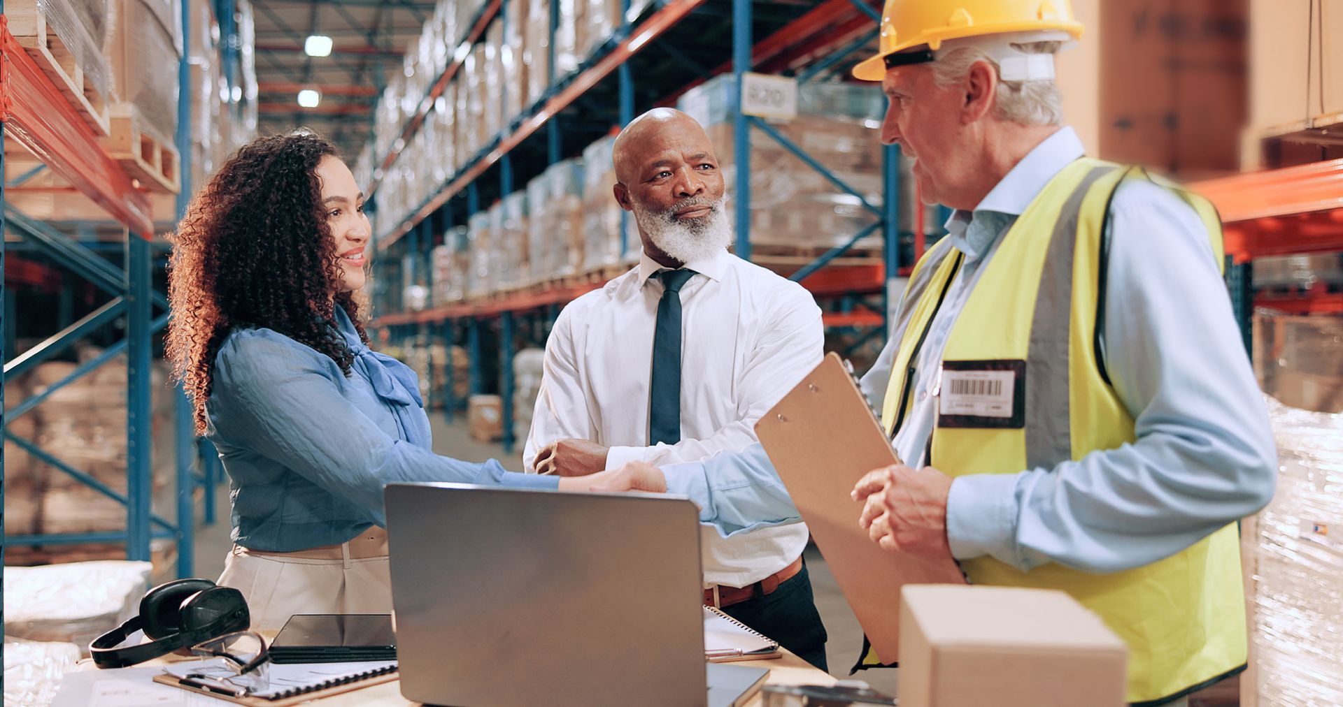 Three people in a warehouse discuss a shipment beside boxes and a laptop.