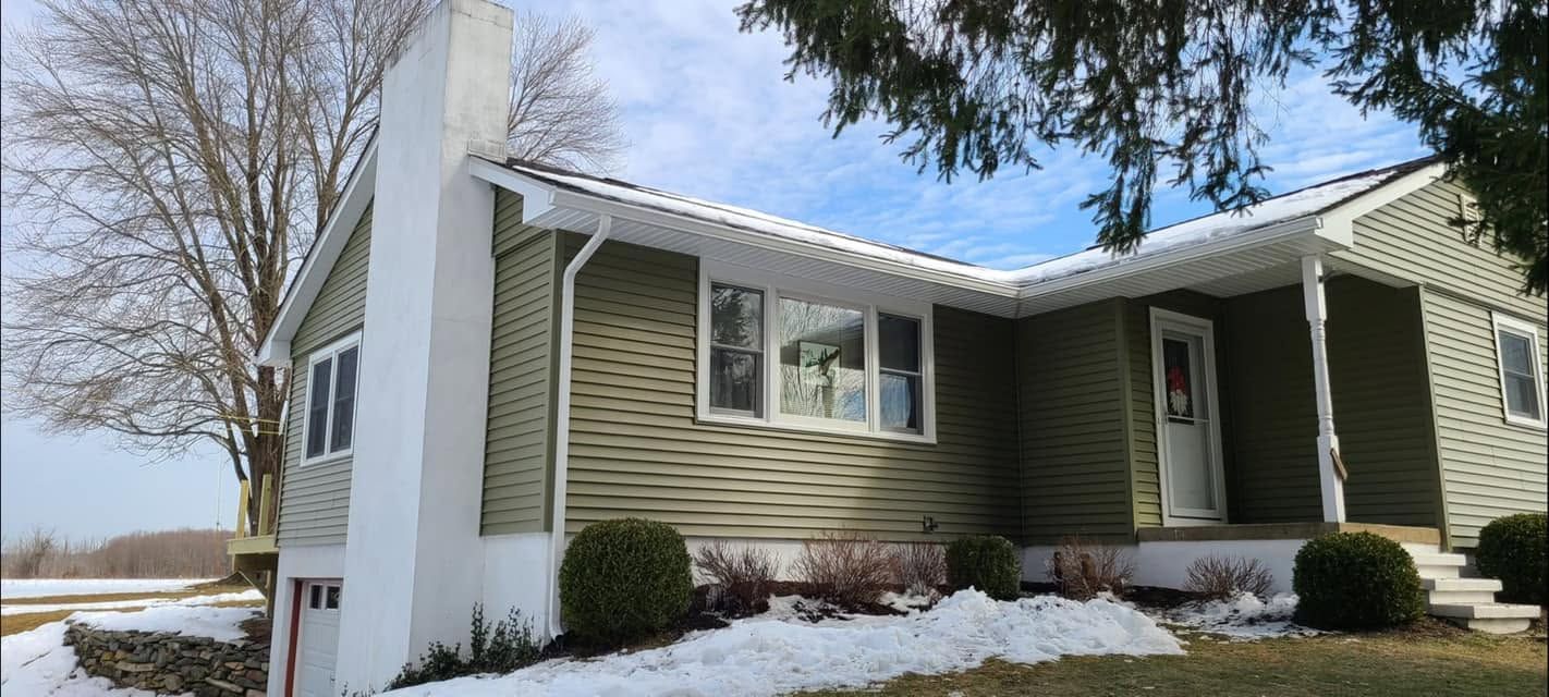 Snowy suburban house with green siding, white trim, and a white chimney under a blue sky