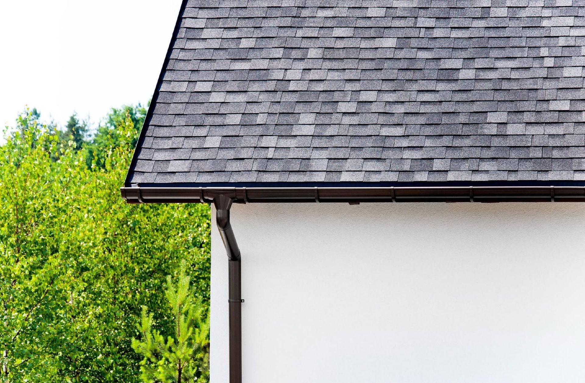House roof with gray shingles and white wall beside green trees