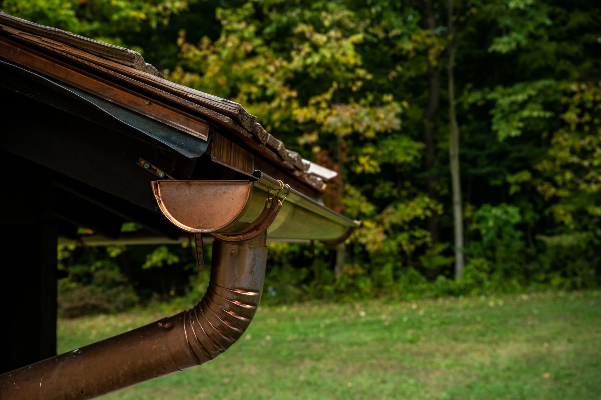 Close-up of a brown rain gutter and downspout on a house roof, with green trees and lawn in the background