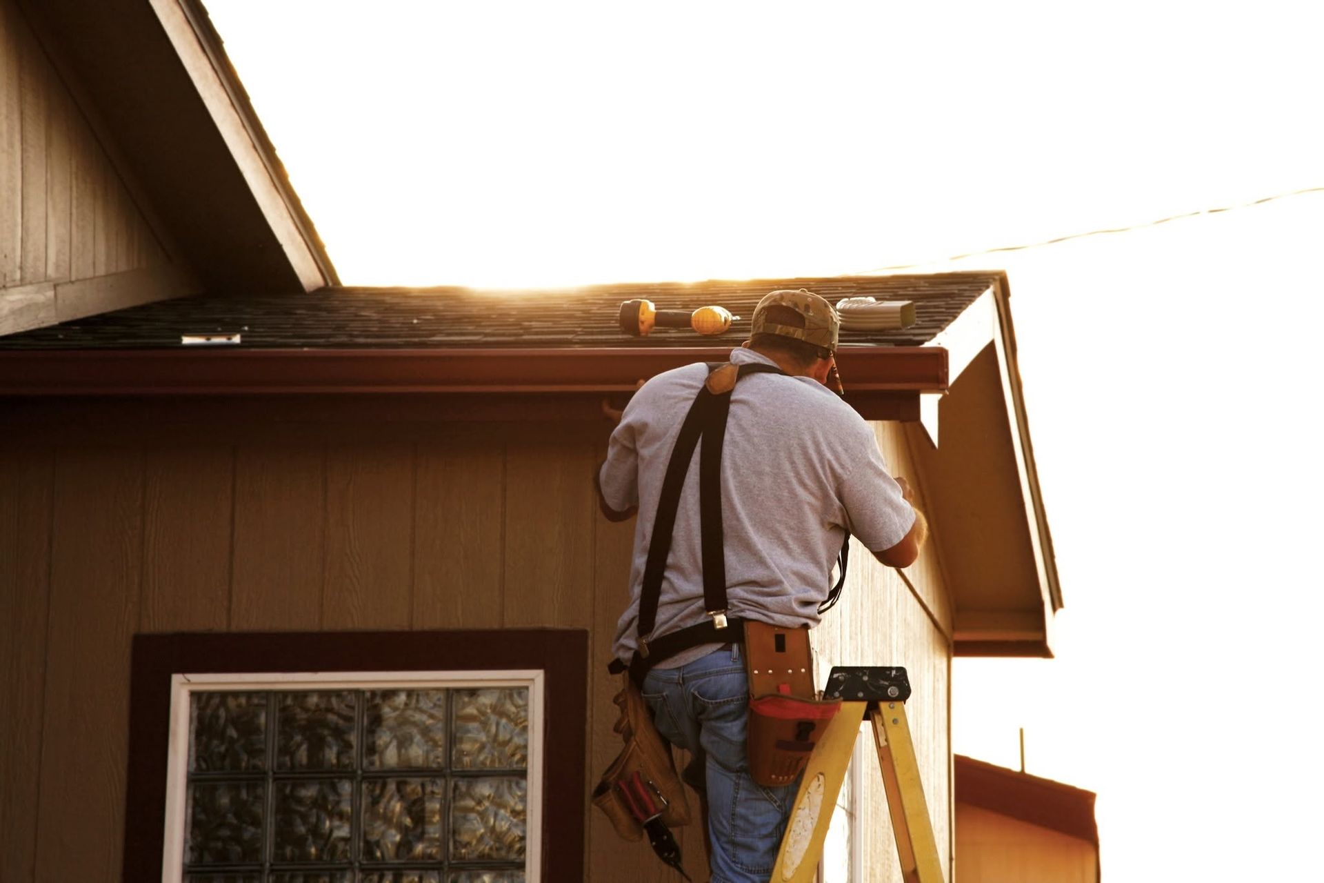 Worker on a ladder repairing the roof edge of a house at sunset