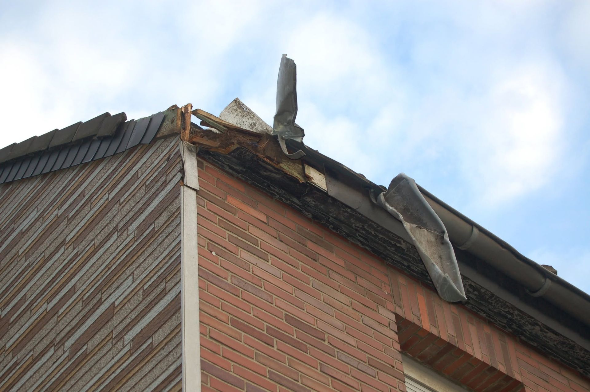 Damaged brick building roof with torn black shingles and debris hanging off the edge under a blue sky