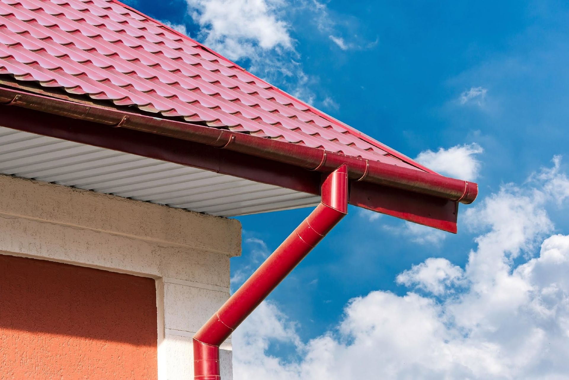Red rain gutter and downspout on a house with a red roof against a blue sky with clouds