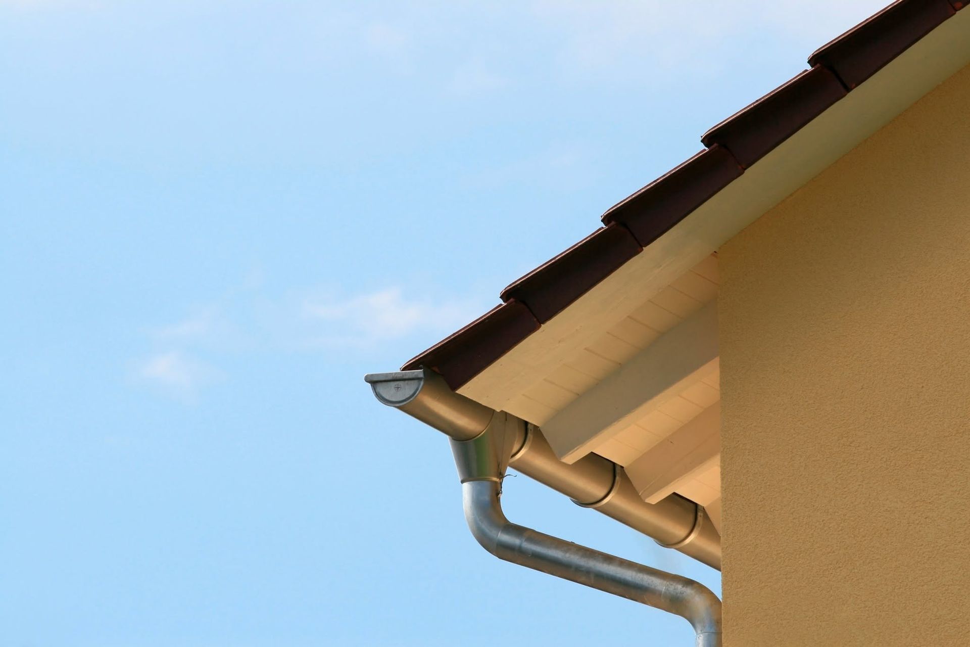 Gutter and downspout on a beige house roof against a blue sky