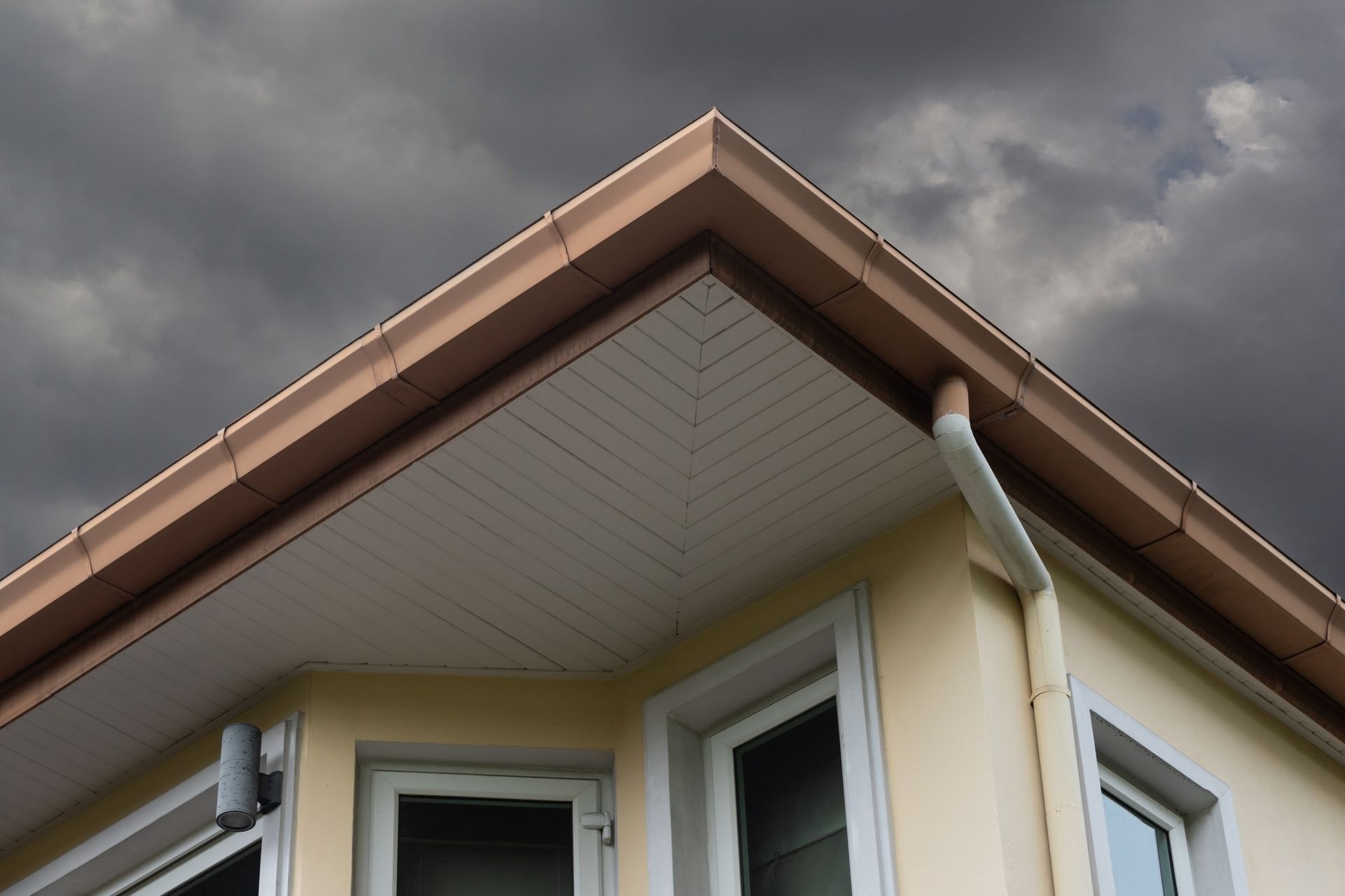 Close-up of a house roof corner with beige siding, brown trim, and two windows below