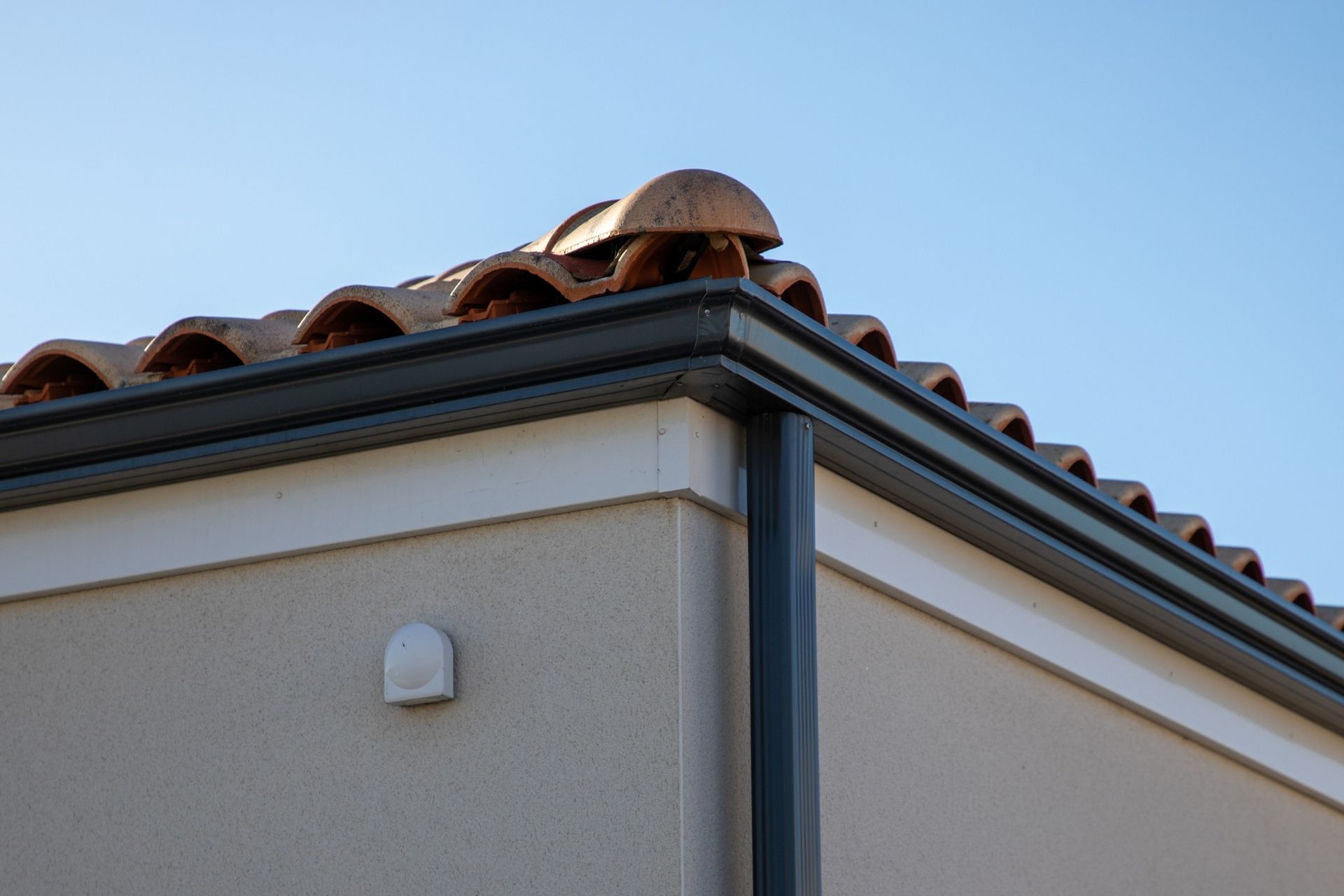 Corner of a stucco house with gray trim and terracotta roof tiles against a blue sky