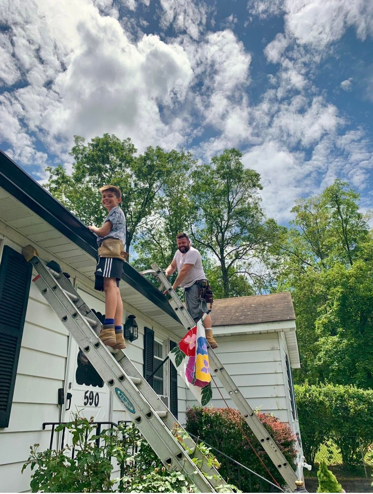 Two workers on a roof with ladders, cleaning gutters beside a white house under a cloudy sky