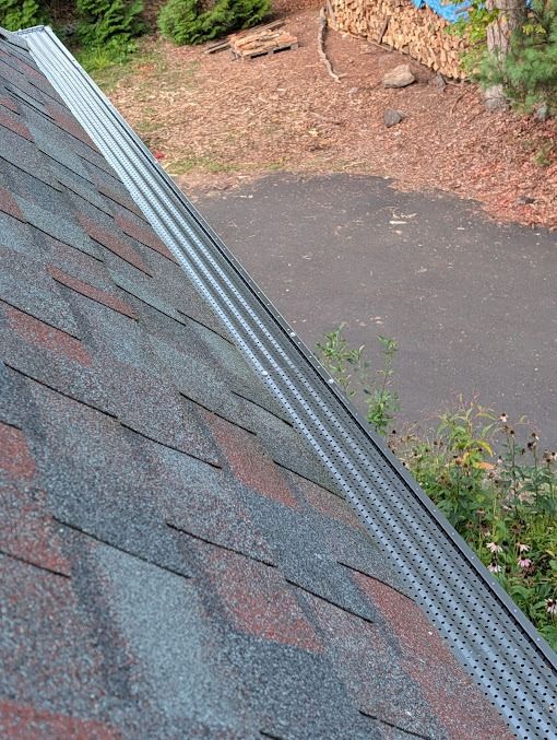 Roof shingles and metal gutter over a driveway with scattered leaves and nearby shrubs