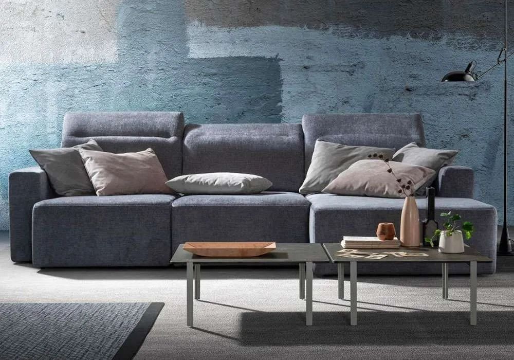 Divano blu con cuscini di fronte a una parete con texture blu e grigia, accanto a due tavolini da caffè quadrati.