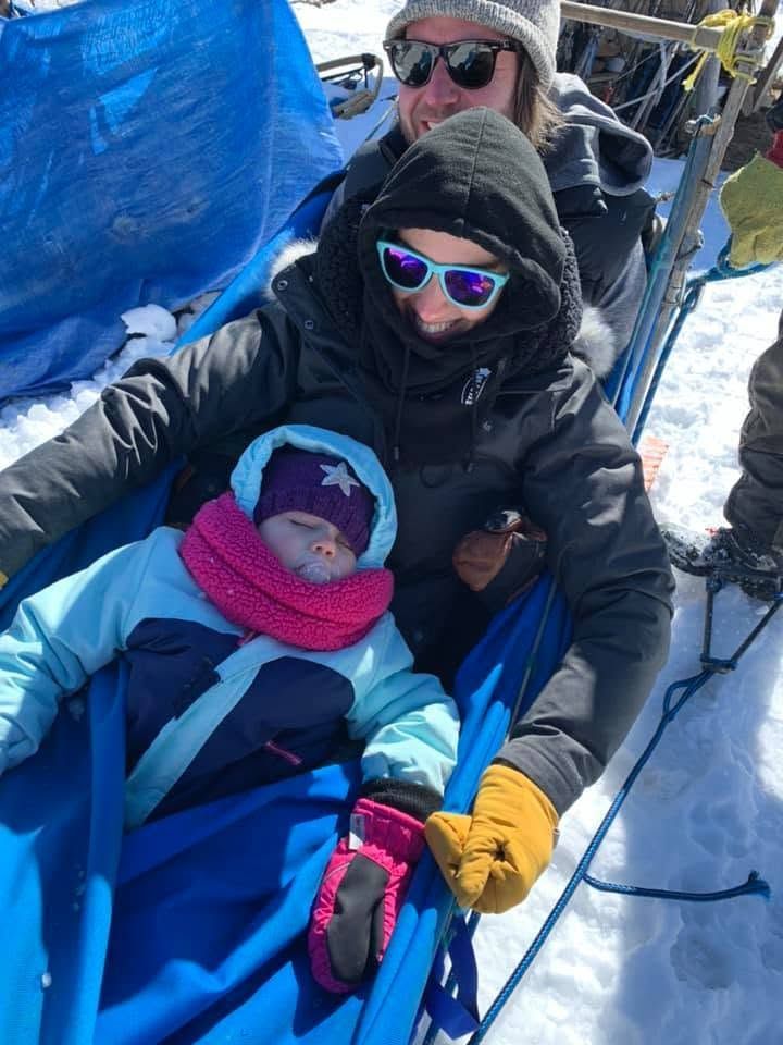 Three bundled people seated in a sled on a snowy mountain, one holding a baby in pink.