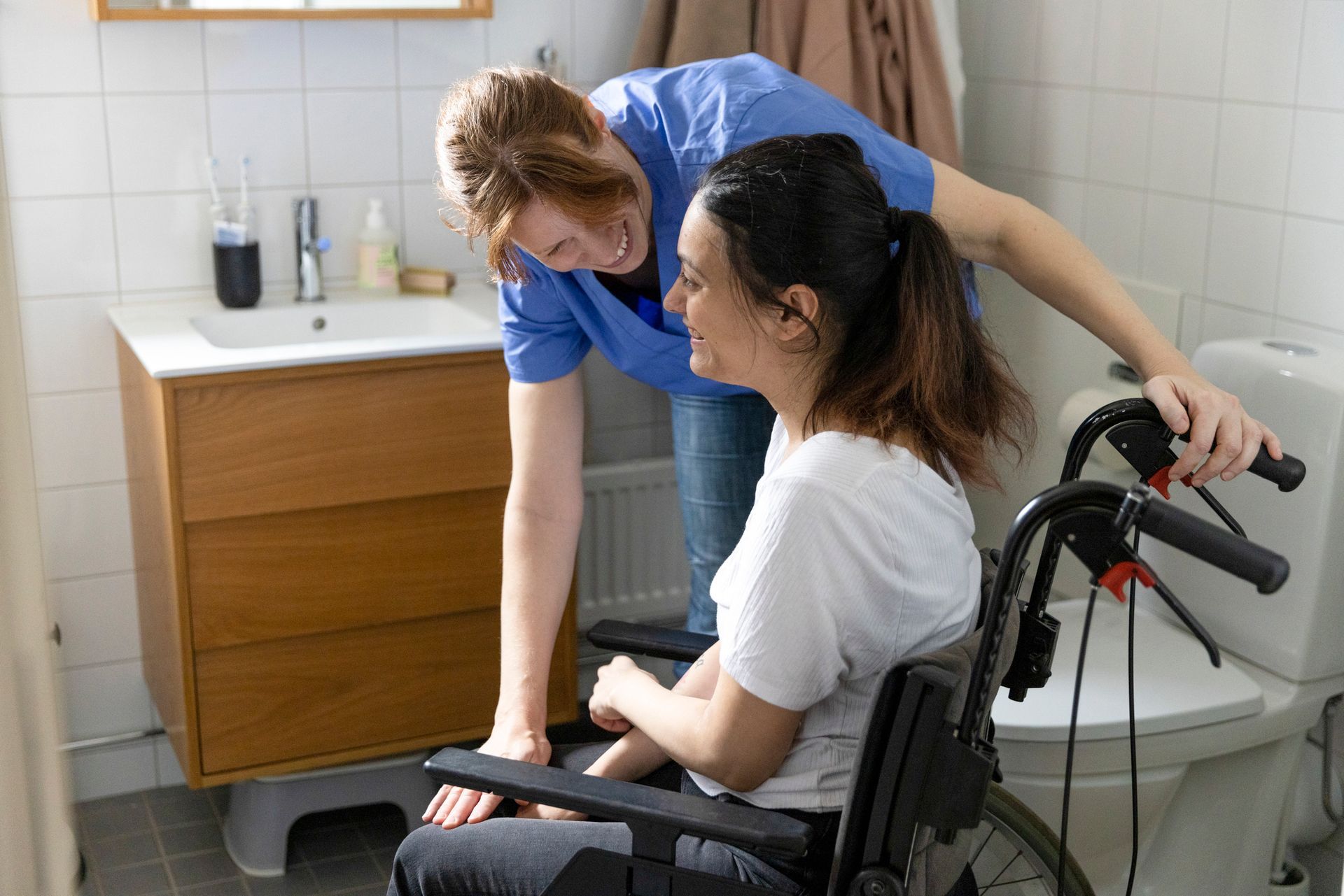 Happy female nurse talking to a woman with paraplegia in a bathroom at home.