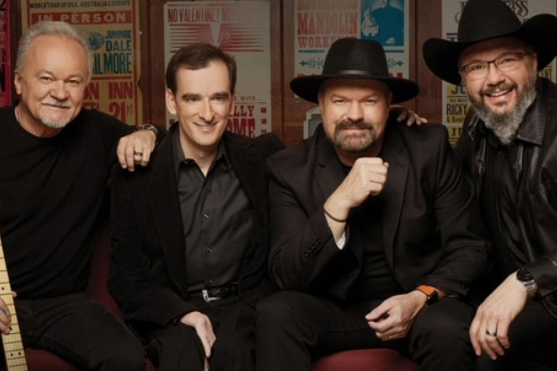 Four men, band members, smiling, posed closely together. Background of vintage signage.