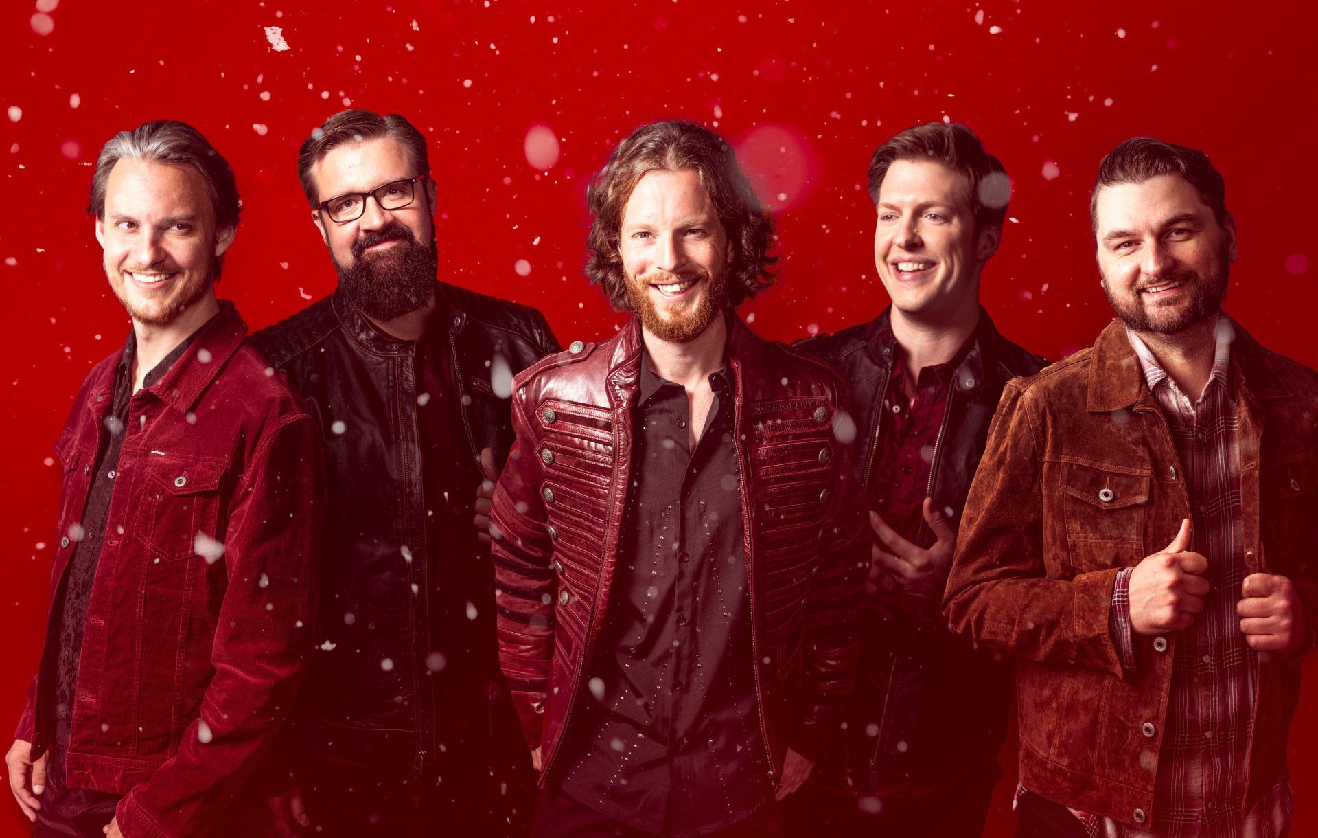 A group of five men smiling and posing against a red background. Snowflakes fall.
