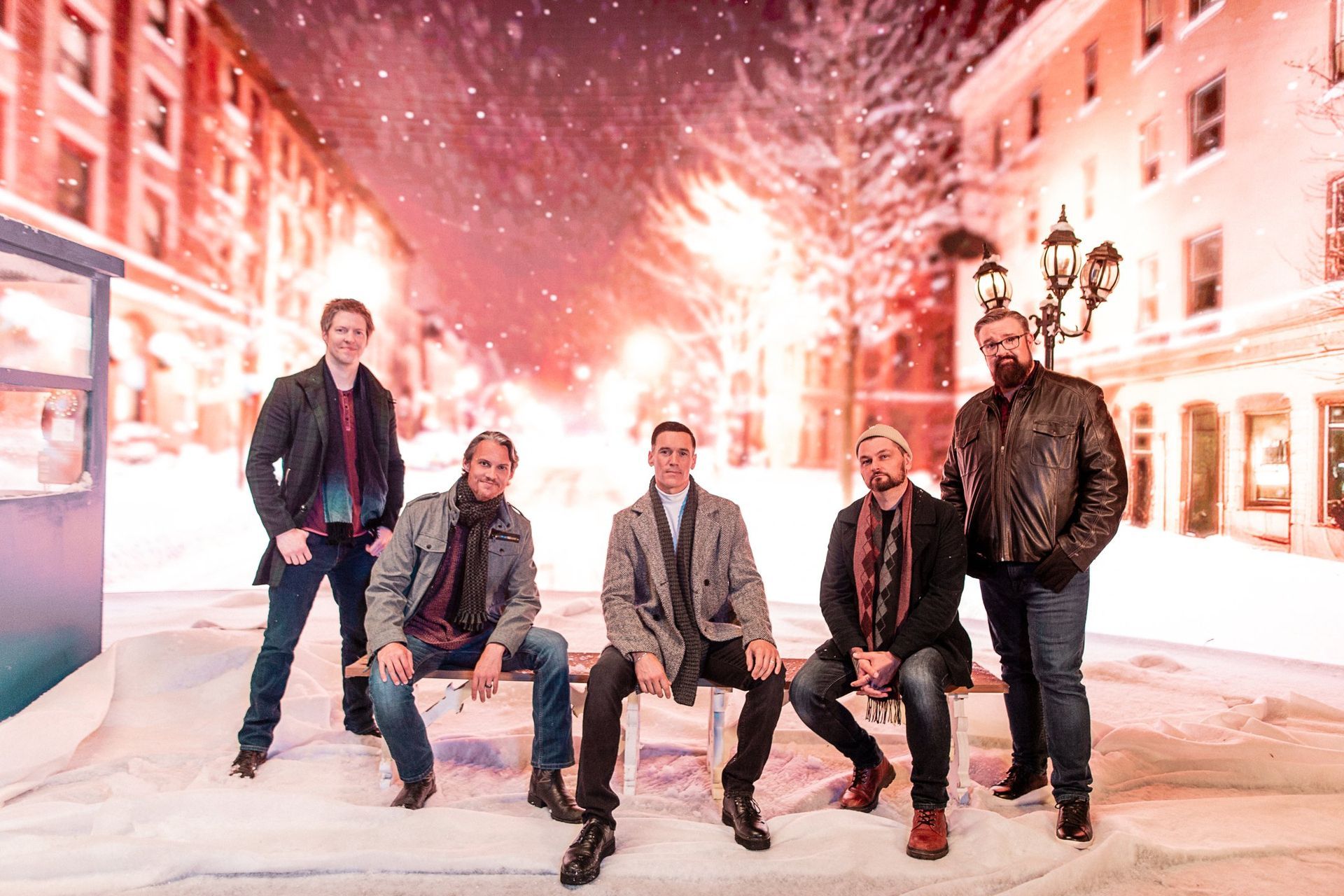 Five men posing on a bench in a snowy street scene, likely a holiday promo.