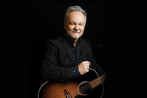 Man holding a guitar, smiling. He is wearing a blazer and button-down shirt with a dark background.