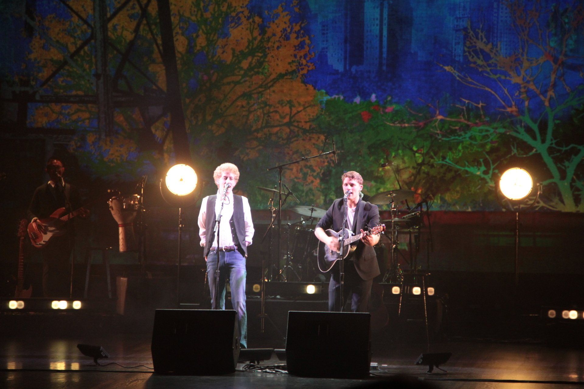 Band performing on stage with backdrop of trees. Two singers center, guitarist right, drummer and bassist left.