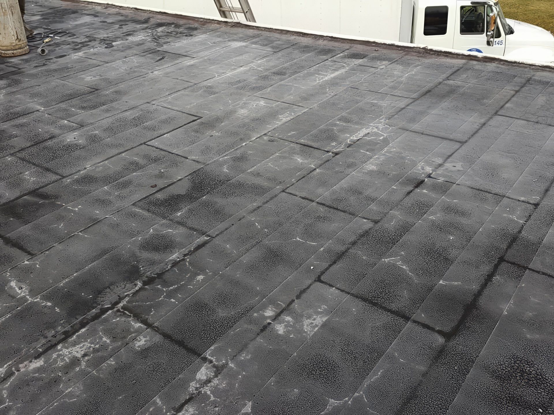A wide, grey, flat commercial roof with moisture patterns, a yellow tool, and a beige wall under a cloudy sky.