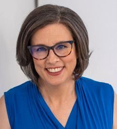 Smiling woman with short brown hair and blue glasses in a blue top against a light background