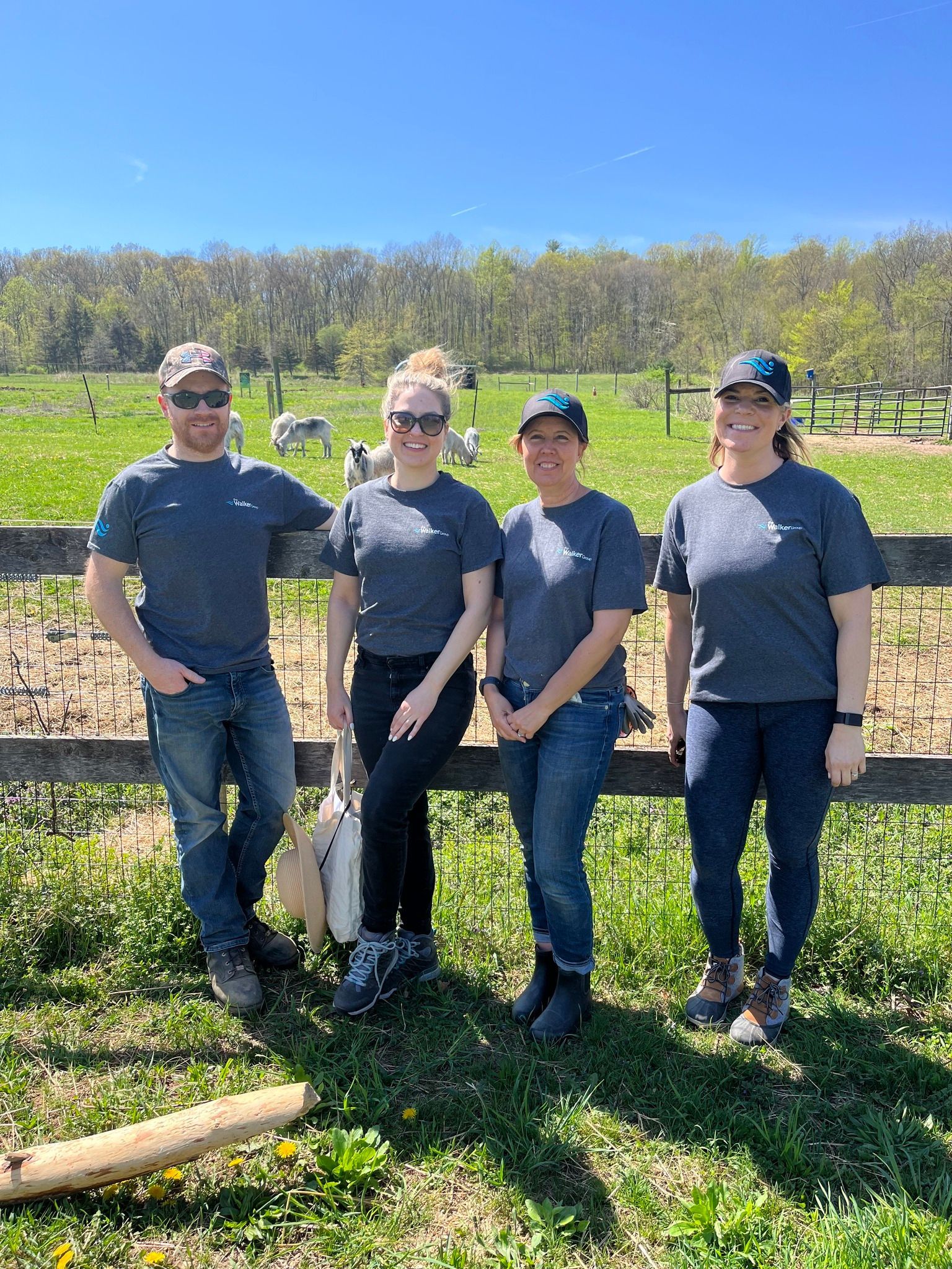 Four people standing in a vineyard on a sunny day, wearing casual gray shirts and caps.