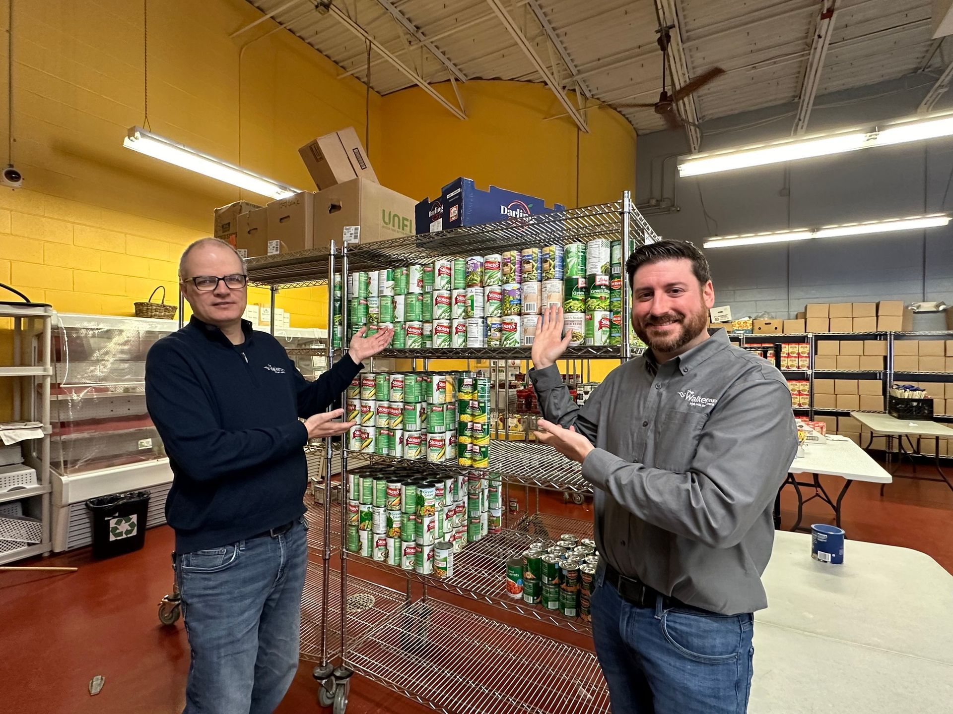 Two men pose beside a tall tower of stacked canned goods in a warehouse.