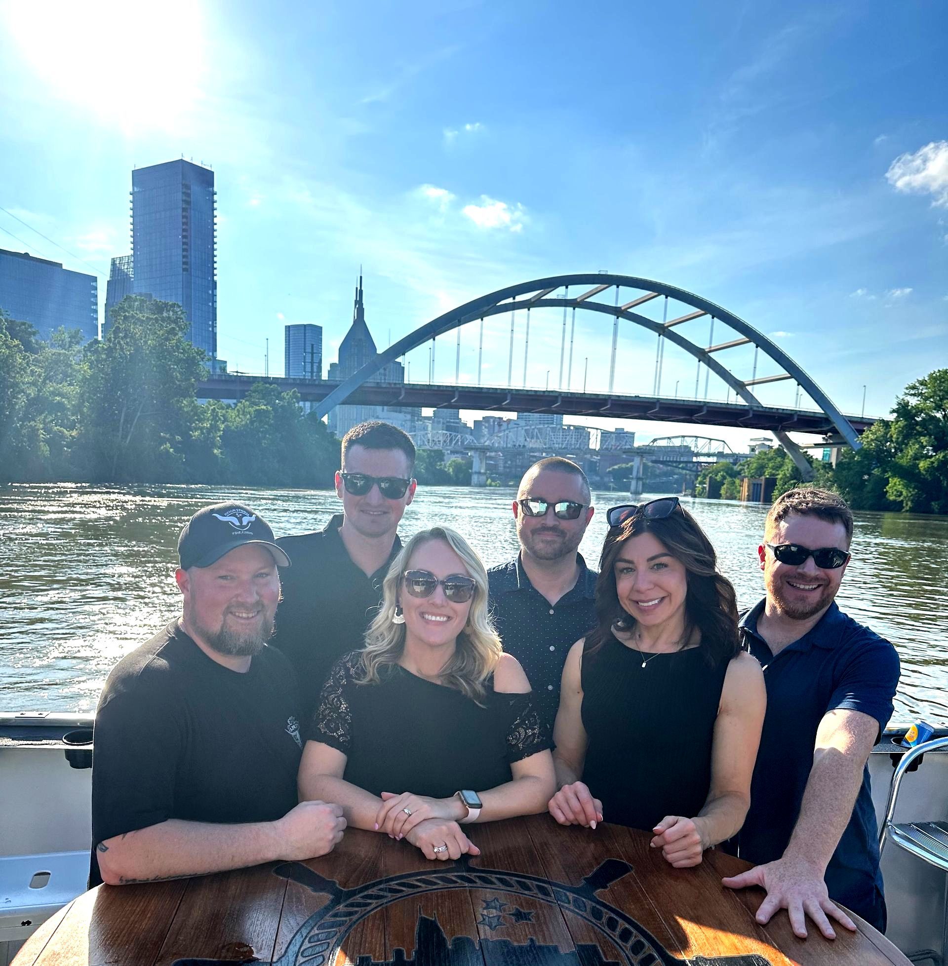 Six friends smiling by a river with a large arched bridge and city skyline behind them.