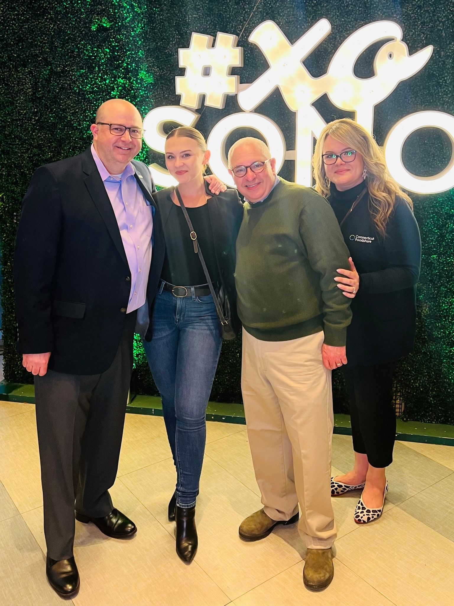 Four people posing indoors in front of a lit “#XGO” sign