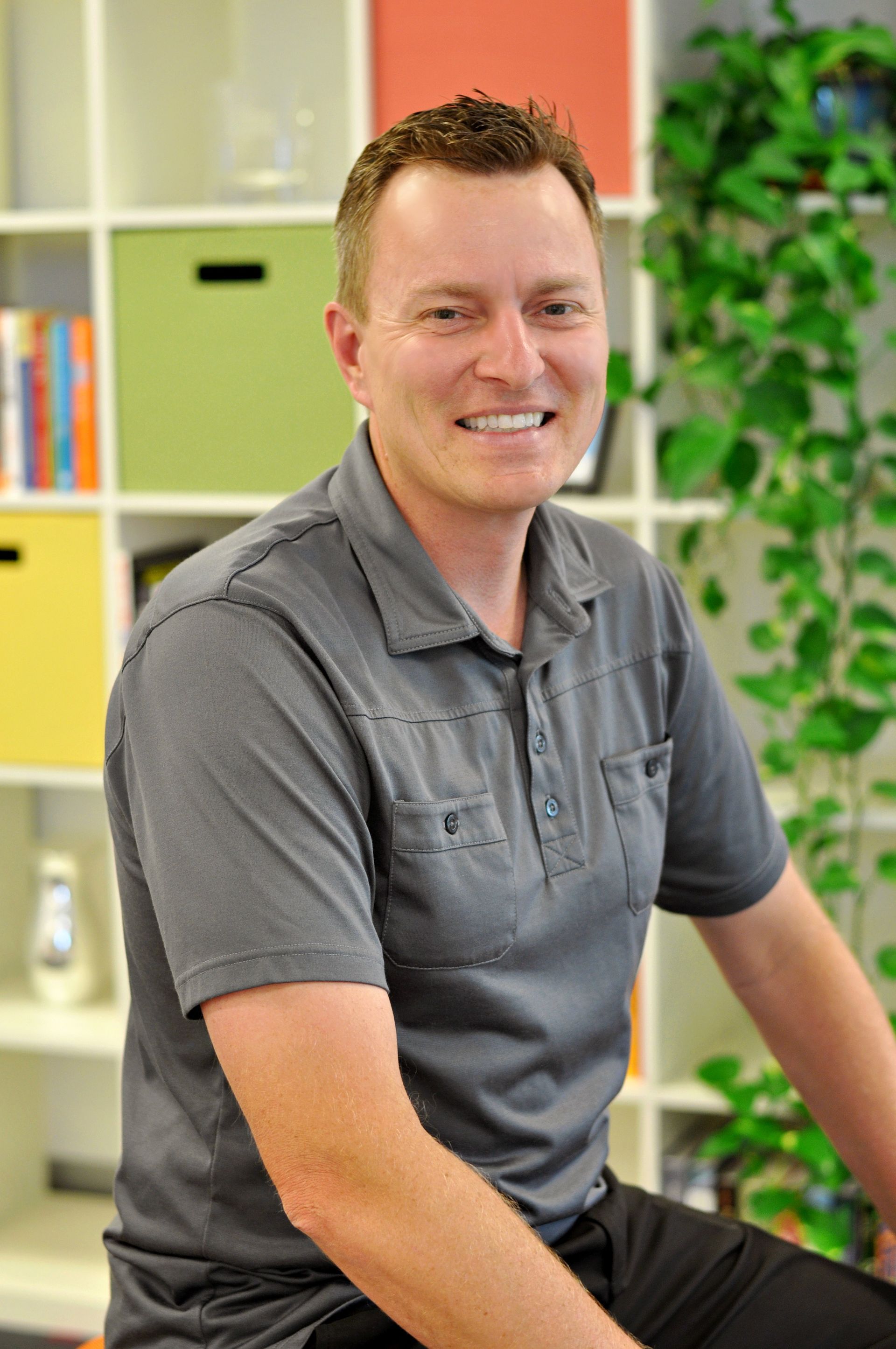 Smiling man in a gray shirt sitting indoors beside colorful shelves and a green plant