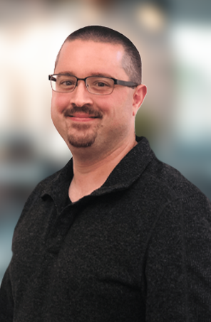 Portrait of a smiling person wearing glasses and a dark sweater against a blurred background
