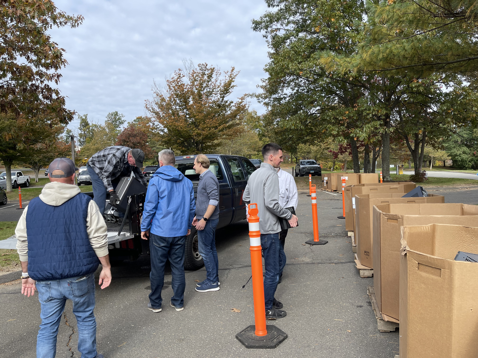 People standing by an open truck near rows of cardboard boxes in a parking lot